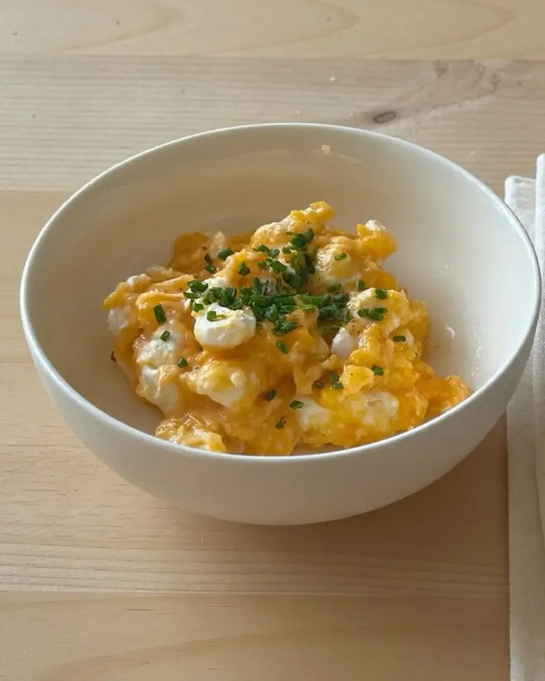A white bowl of scrambled eggs with cottage cheese curds, topped with chives, on a wooden table.