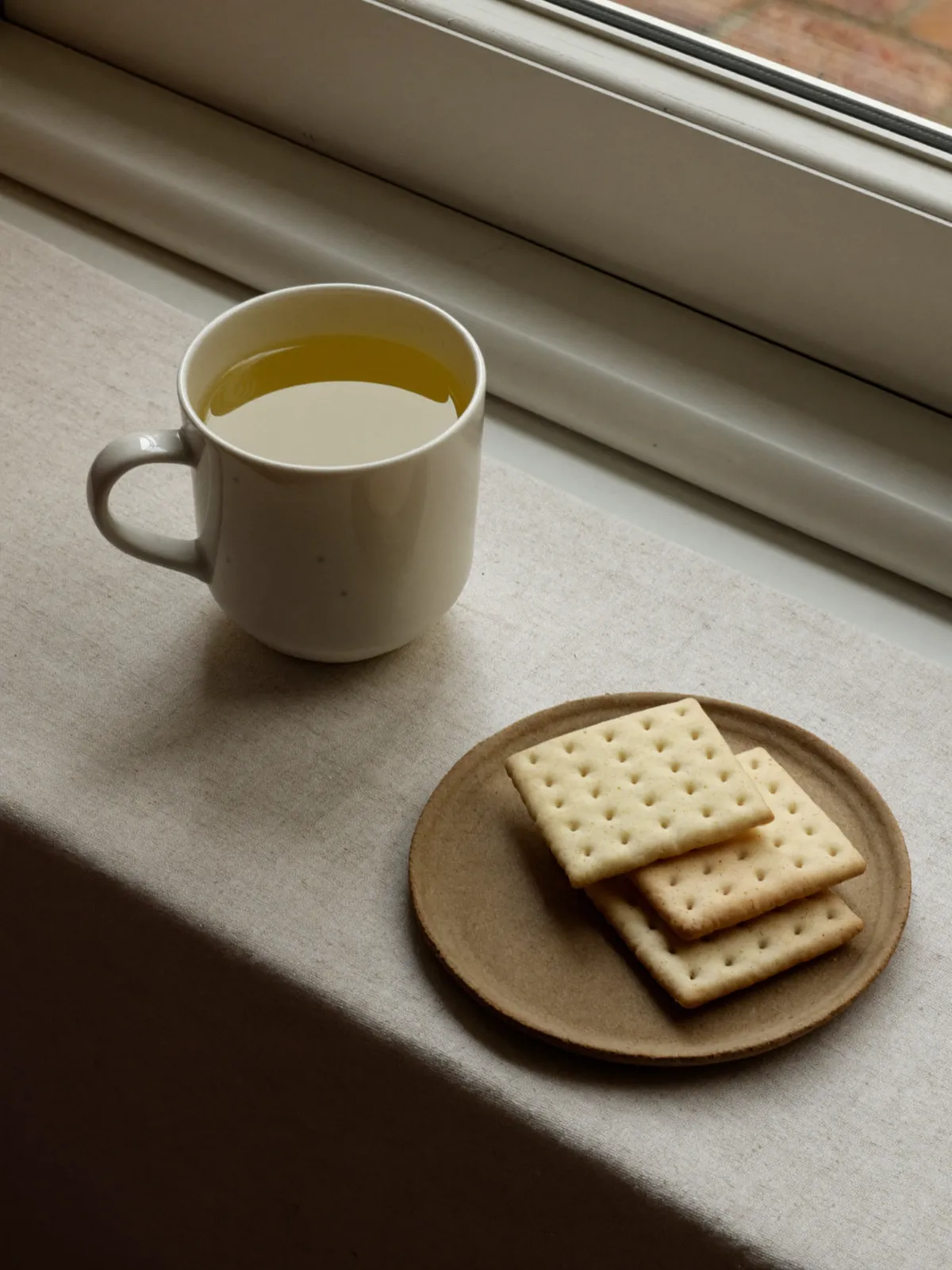 A cup of pale ginger tea and a small plate of plain crackers on a windowsill table.
